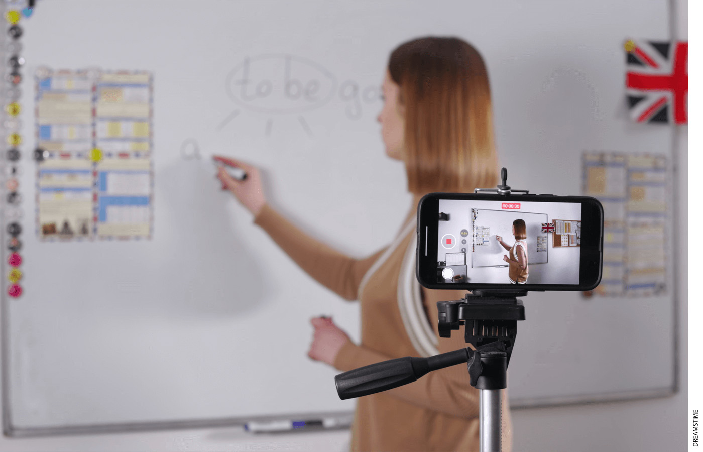 Photo of a teacher writing on a white board while being filmed on a phone