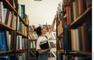 A student in a library reaches up to the top shelf for a book.