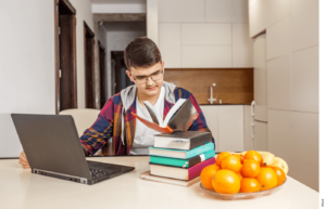 A student sits at a table with a laptop and a pile of books