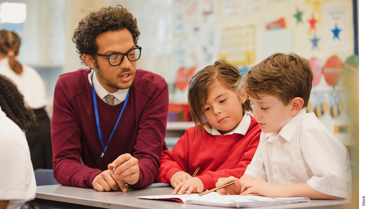 A teacher instructs two students at a classroom table.