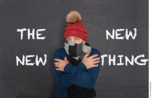 A person wrapped in a scarf and winter hat shivering standing in front of a chalkboard with "THE NEW NEW THING" written on it.