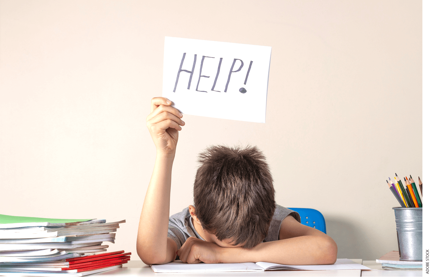 Boy student with head down on desk holding up a sign that says "Help!" Pile of notebooks to his right, container of colored pencils to his right.